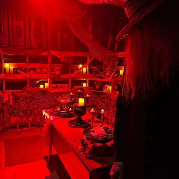 Spooky room bathed in red light, featuring candles, skulls on shelves, cobwebs, and a figure wearing a hat